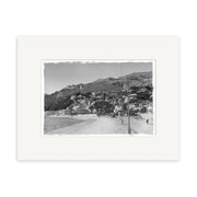 The Oriental Bay 1900s - HI009 is a black and white historical photographic print featuring a coastal town with hillside houses, sandy beach, and people strolling by the shore, framed with a white mat and printed on archival matte paper.