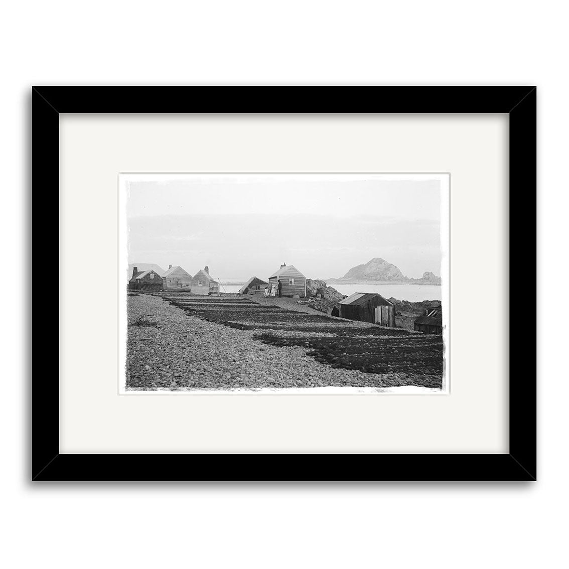 Island Bay 1900 No.1 - HI010: Black-and-white historical photo of wooden houses by a rocky shore, hills, and calm water, printed on archival matte paper in a black frame with white mat.