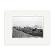 Island Bay 1900 No.1 - HI010: Black and white historical photographic print of small wooden buildings on a rocky shore with mountains in the distance, expertly framed with a wide white mat on archival quality matte paper.