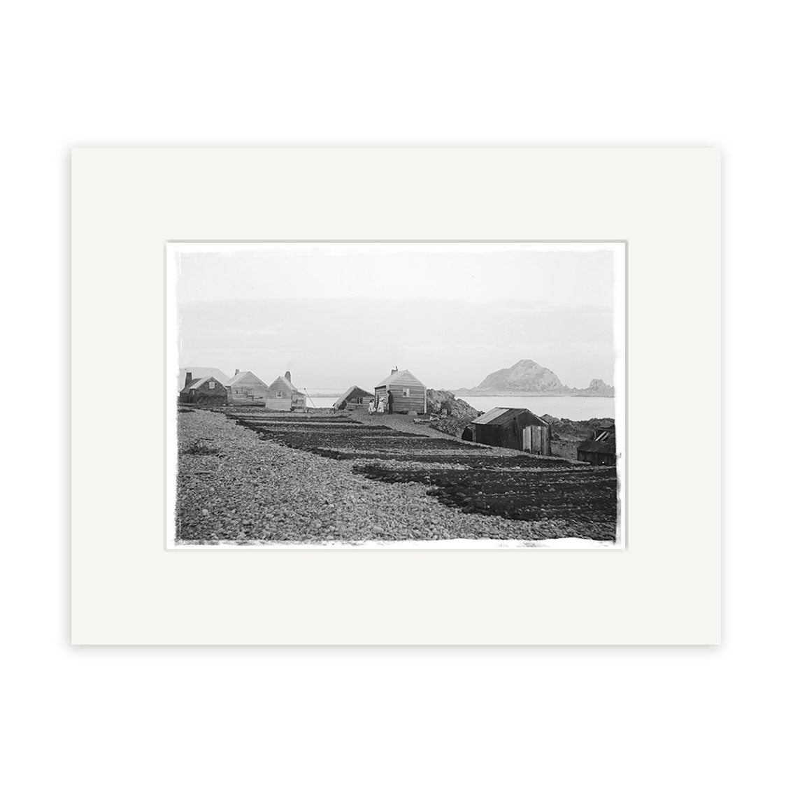 Island Bay 1900 No.1 - HI010: Black and white historical photographic print of small wooden buildings on a rocky shore with mountains in the distance, expertly framed with a wide white mat on archival quality matte paper.