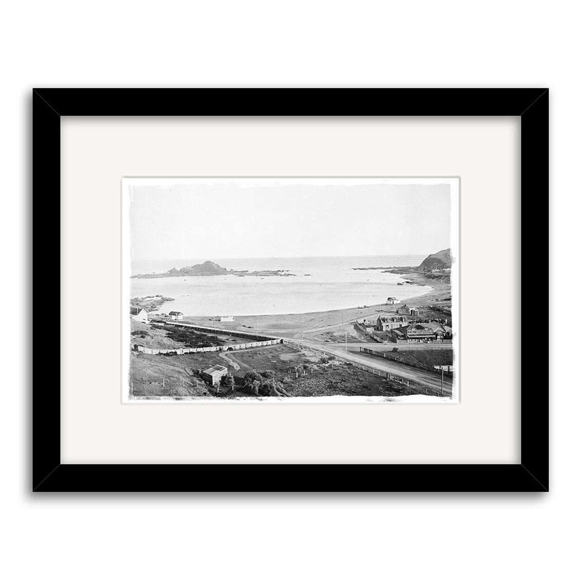 Island Bay 1900 No.2 - HI011: Black-and-white historical photo print of a coastal landscape with buildings, beach, and rocky islands, presented in a black frame with white mat on matte paper.