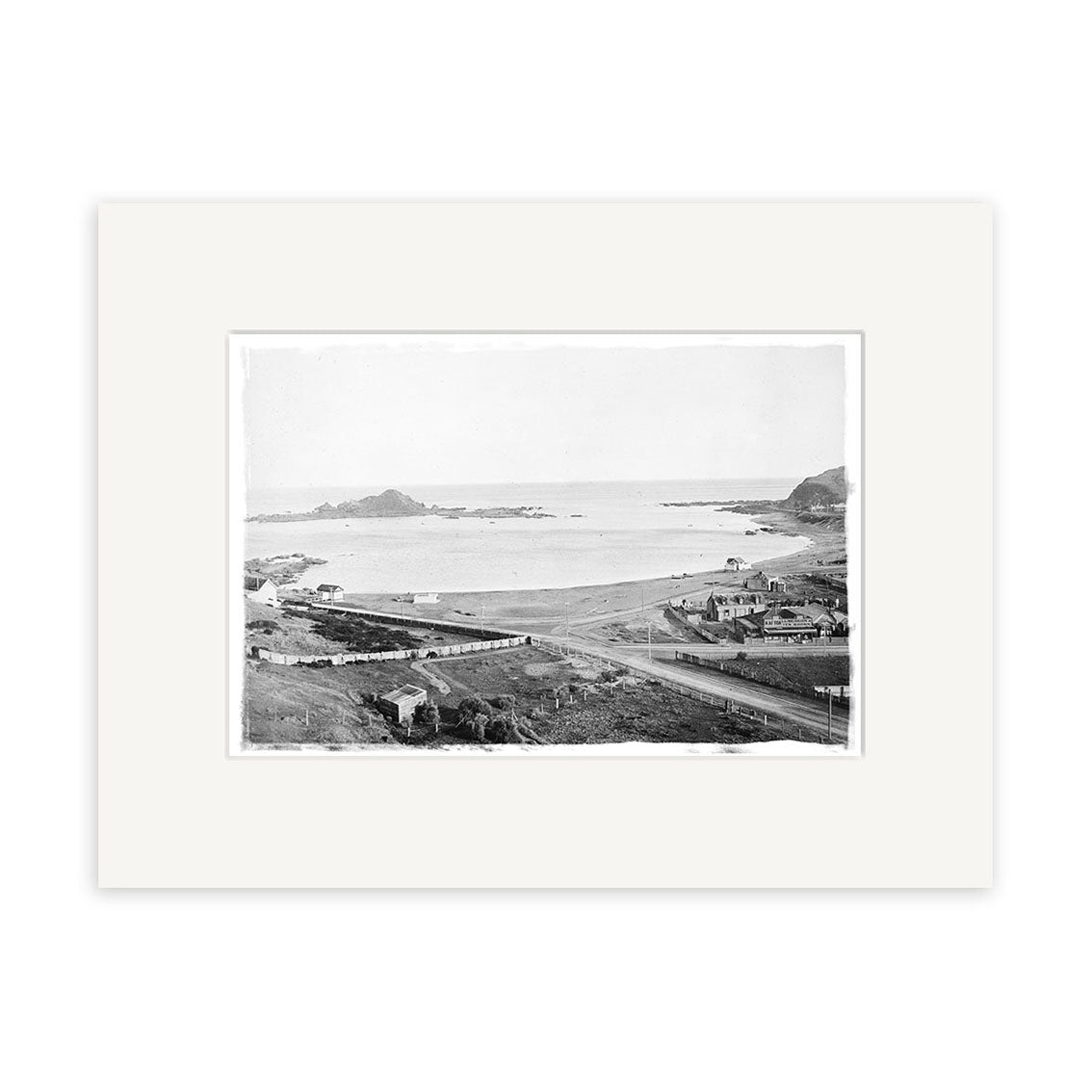 Island Bay 1900 No.2 - HI011 is a black and white historical photographic print depicting a bay, rocky shoreline, and houses. Matted with a white border on matte paper for archival quality preservation.
