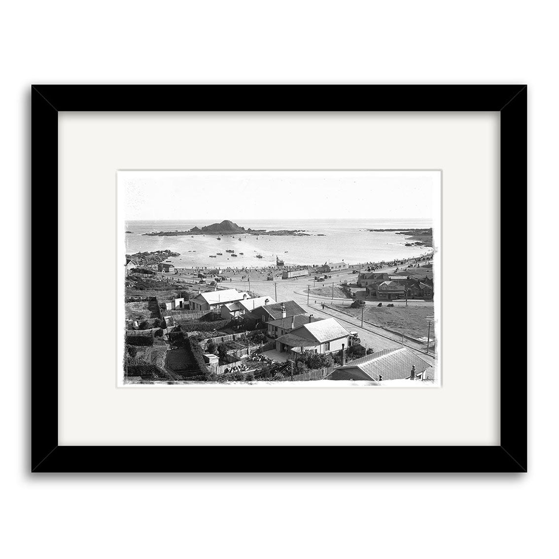 Island Bay 1920s - HI012: Black and white photo of a New Zealand coastal town with houses, roads, and a busy beach. This historical print is framed with a white mat and black border for archival quality.