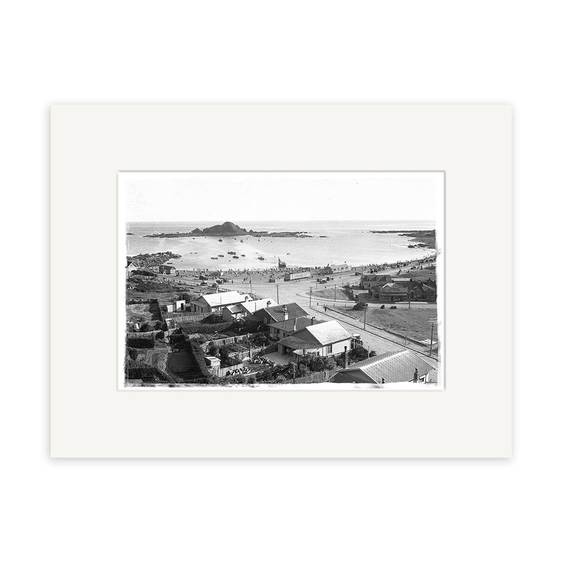 Black and white photo, Island Bay 1920s - HI012, shows a New Zealand coastal town with houses, roads, people on a sandy beach, ocean, and rocky islands. This historical photographic print includes a white mat border.