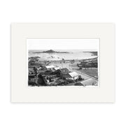 Black and white photo, Island Bay 1920s - HI012, shows a New Zealand coastal town with houses, roads, people on a sandy beach, ocean, and rocky islands. This historical photographic print includes a white mat border.