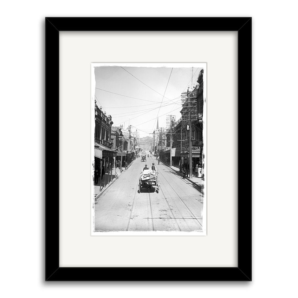 Willis St 1900 - HI013: Black and white photo print of an early 20th-century city street with a streetcar, horse-drawn wagons, buildings, and scaffolding. Framed in black with a white mat on archival quality matte paper.