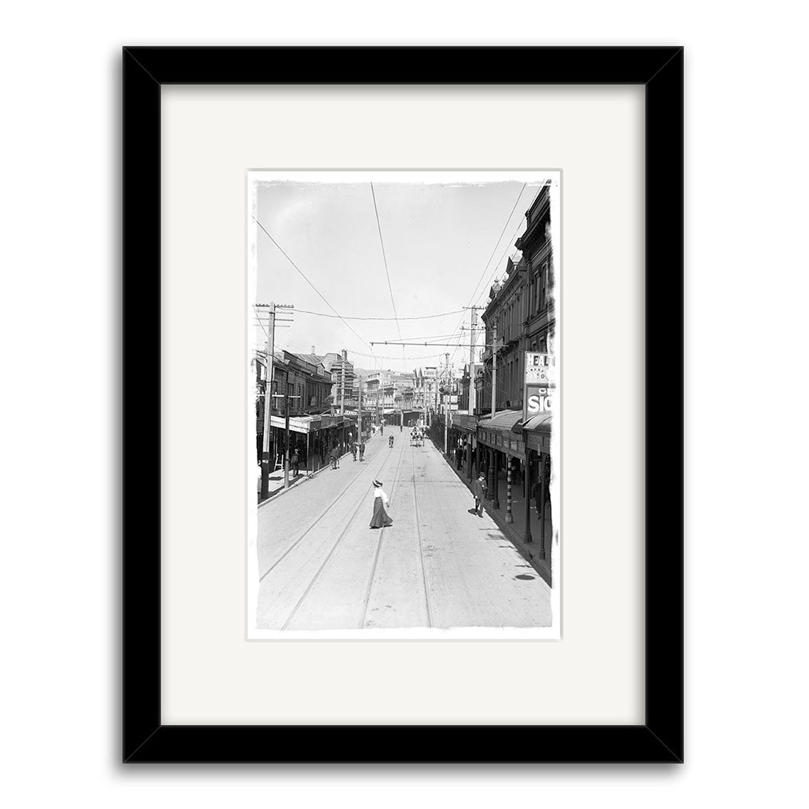 Manners St 1907 - HI014: Black-and-white city street photo with shopfronts, pedestrians, and trolley wires. A woman stands in the middle. Framed in black with white mat on archival quality matte paper.