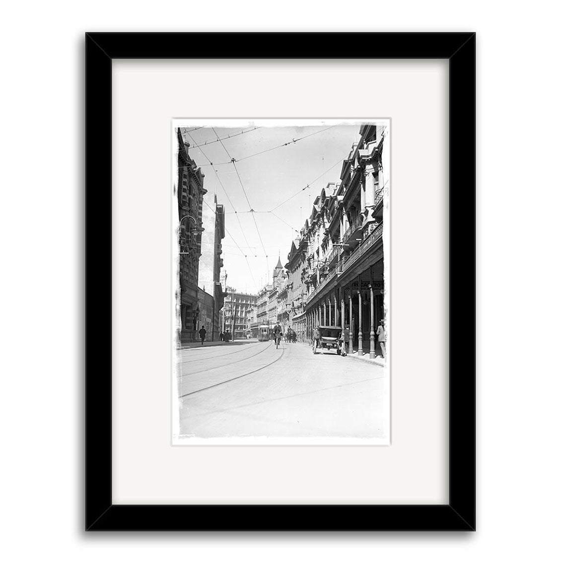 Framed black-and-white photo print, Lambton Quay 1900 - HI015, shows a historic New Zealand city street with old buildings, tram wires, columns, cars, and pedestrians. Printed on archival quality paper for lasting preservation.