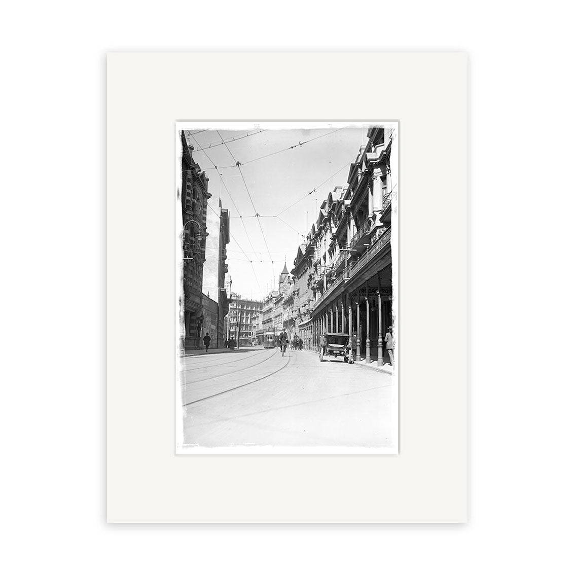 Lambton Quay 1900 - HI015: Black and white historical photographic print of an old New Zealand city street with tram tracks, early cars, pedestrians, and buildings with columns and balconies. Overhead wires span this archival quality scene.