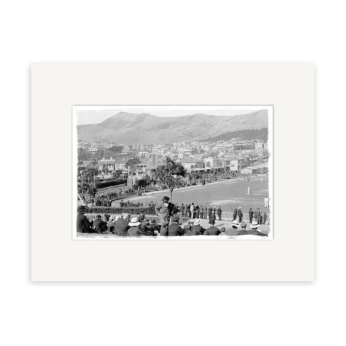 Black and white photo, Basin Reserve 1900 - HI016: cricket match in progress with players on the field, spectators in hats, buildings and hills behind. Archival quality matte paper, made in New Zealand.