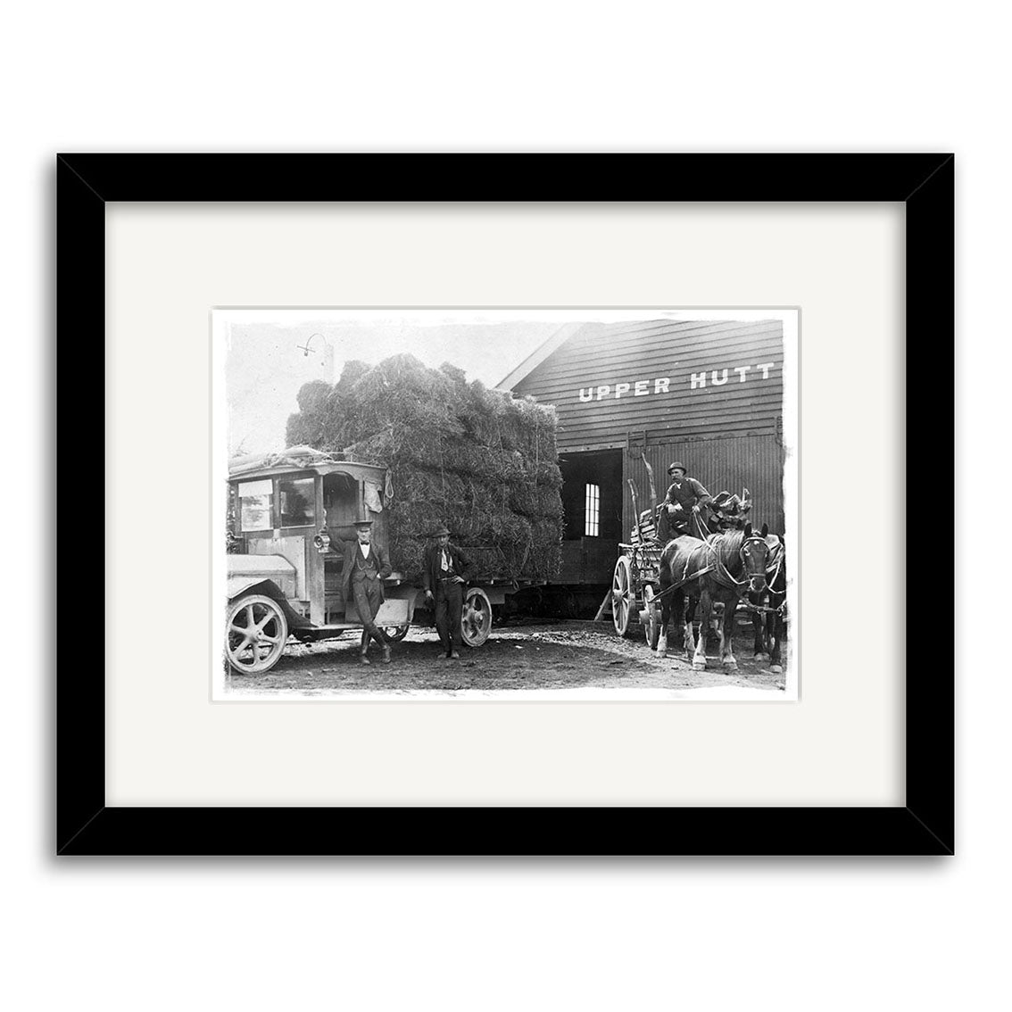 Black and white photo, Upper Hutt 1920 - HI017: A truck loaded with hay is parked by a building labeled Upper Hutt; two men stand beside the truck, near a horse-drawn cart with two horses. Made in New Zealand.