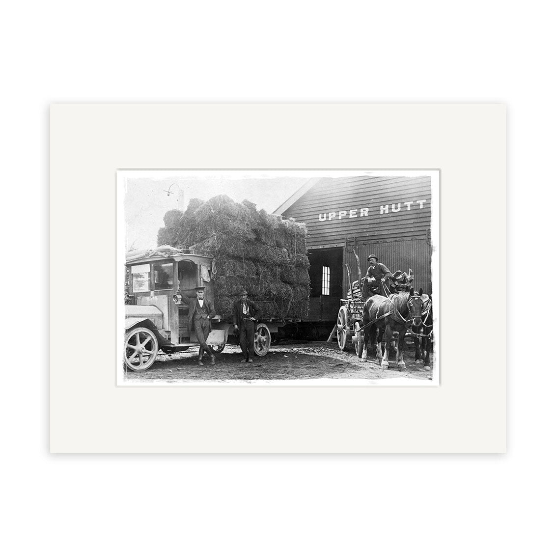 Archival black-and-white photo, Upper Hutt 1920 - HI017: a truck with hay bales and a horse-drawn cart outside an Upper Hutt building; two men by the truck, one on the cart with two horses. Made in New Zealand.