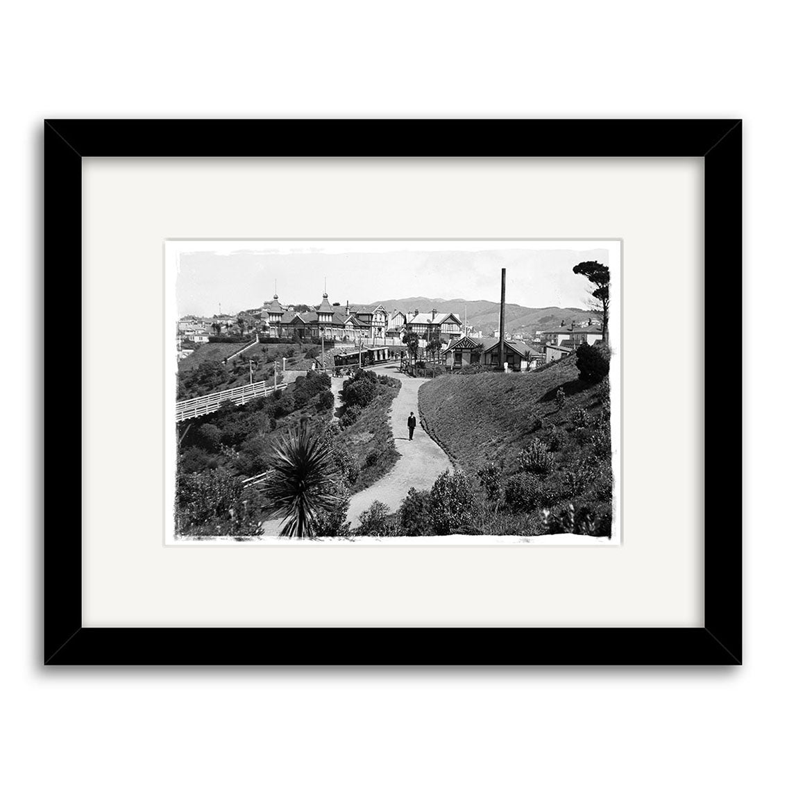 Black and white historical photographic print, Kelburn Car 1918 - HI018, features a winding pathway with a person walking, lush vegetation, and Victorian houses. Printed on archival matte paper, framed with a white mat and black frame.