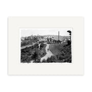 Kelburn Car 1918 - HI018: Black and white photo featuring a vintage car on a hilly road, surrounded by lush plants and Victorian houses, expertly reproduced on archival-quality matte paper.
