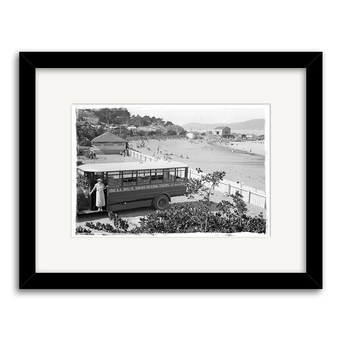 Archival black and white historical photographic print, “Worser Bay 1934 - HI019,” shows a woman by a “Seal Bus” on a sightseeing tour in New Zealand, with beach, ocean, and people in the background. Framed with white mat and black frame.