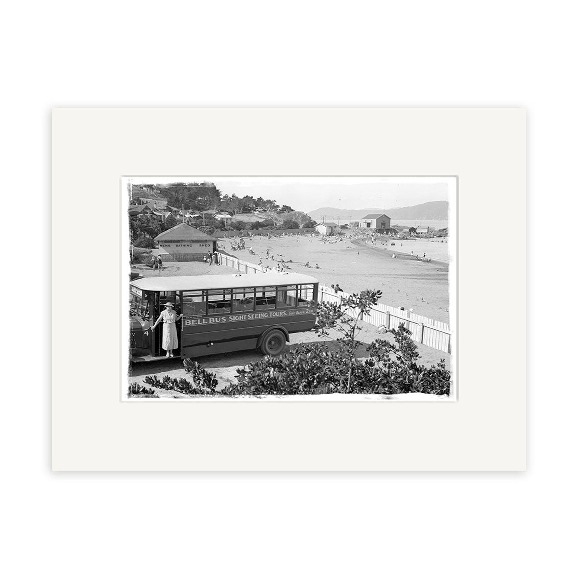 Archival quality black and white photo, “Worser Bay 1934 - HI019”: a vintage Seal Bus, Shark Seeking Tours parked by a New Zealand beach with a woman nearby and people on the sand in the background.