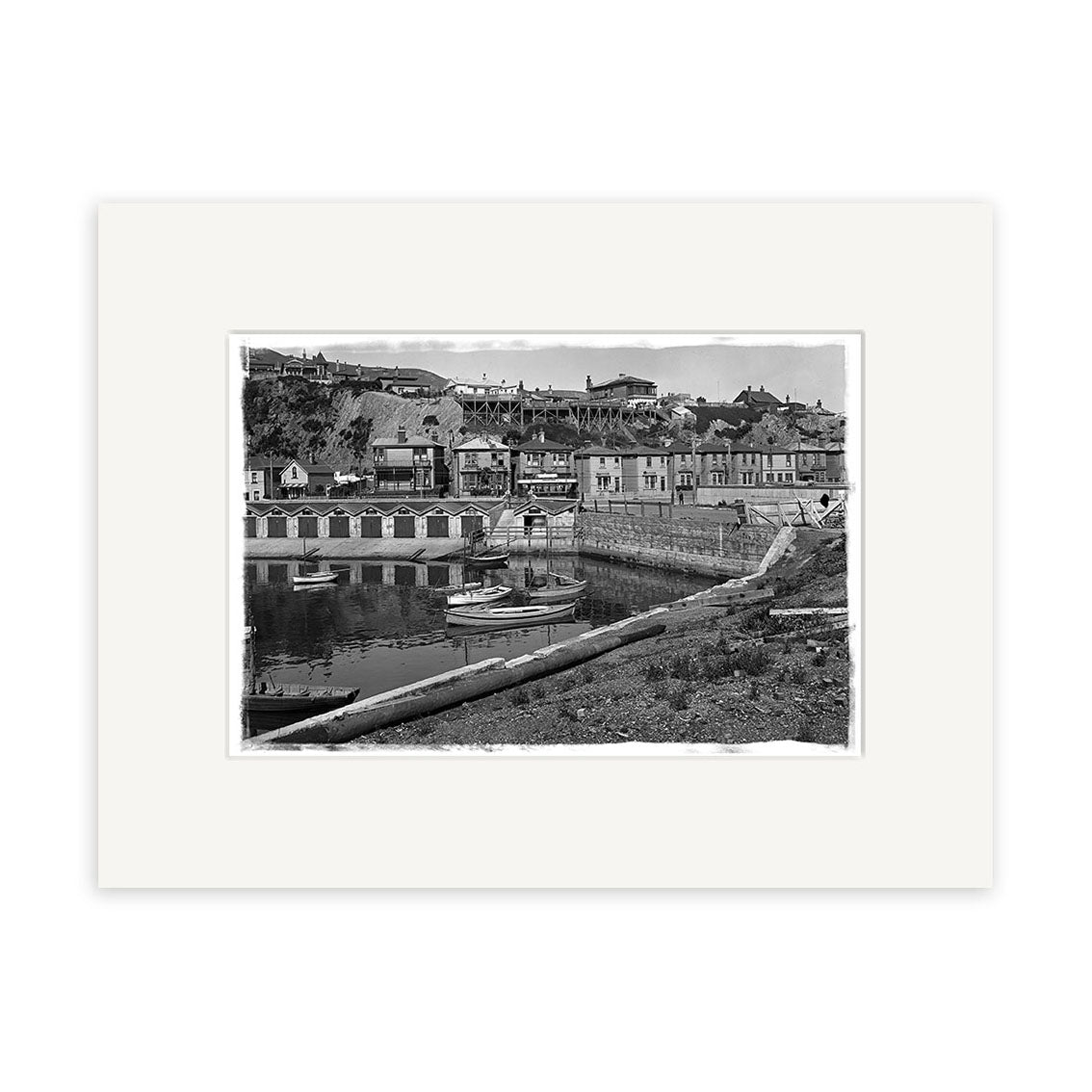 Archival quality black and white photographic print, Oriental Bay 1910 - HI020, features a harbor with small boats, waterfront buildings, and hillside homes; centered in a white mat frame. Made in New Zealand.