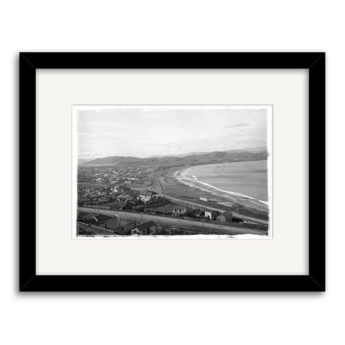 Framed black and white photographic print, Lyall Bay 1910 - HI021: features a coastal town with buildings, beach, railway, and hills. Presented on archival matte paper with a white mat and simple black frame.