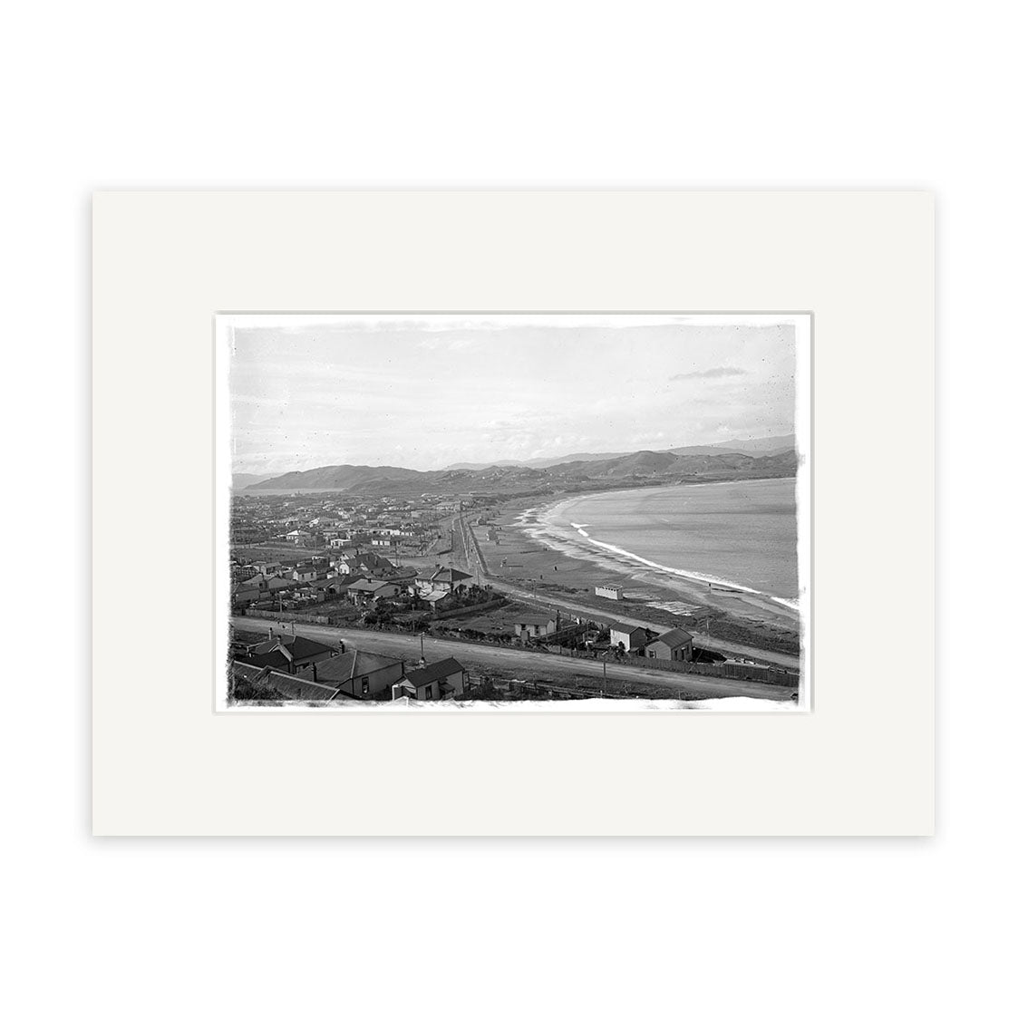 Lyall Bay 1910 - HI021 is a black and white photograph of a coastal town with buildings, a railway line by the shore, and hills behind. This historical print is presented on matte paper to highlight its archival quality.