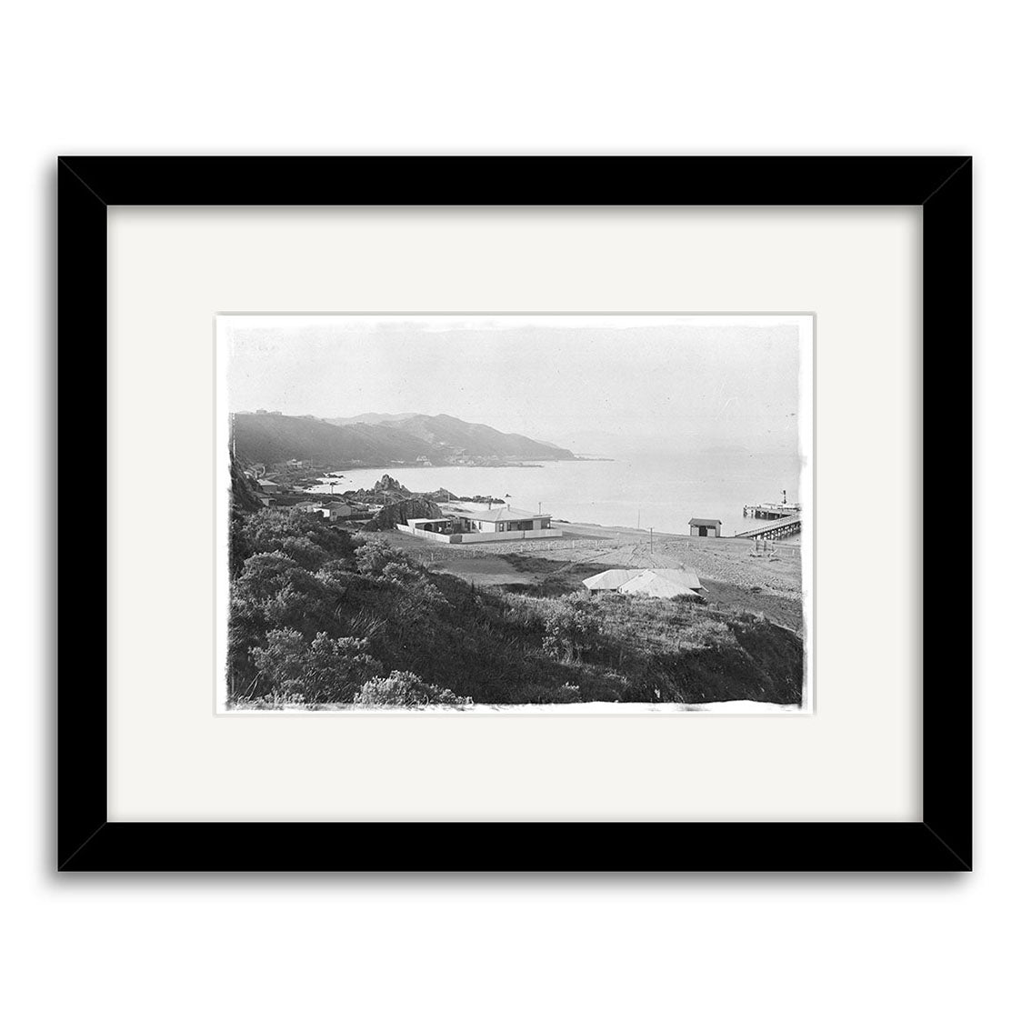 Seatoun 1904 - HI022: Black and white historic photo print of a coastal landscape with buildings, hills, and distant sea. Framed with wide white mat and black frame. Made in New Zealand.