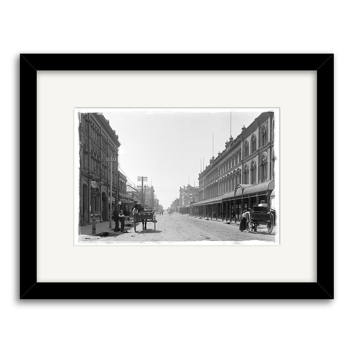 Cashel St 1900 - HI025: Black and white historical photo print of a wide New Zealand city street with multi-story buildings, horse-drawn carriages, and few people, framed in an archival-quality black and white mat.