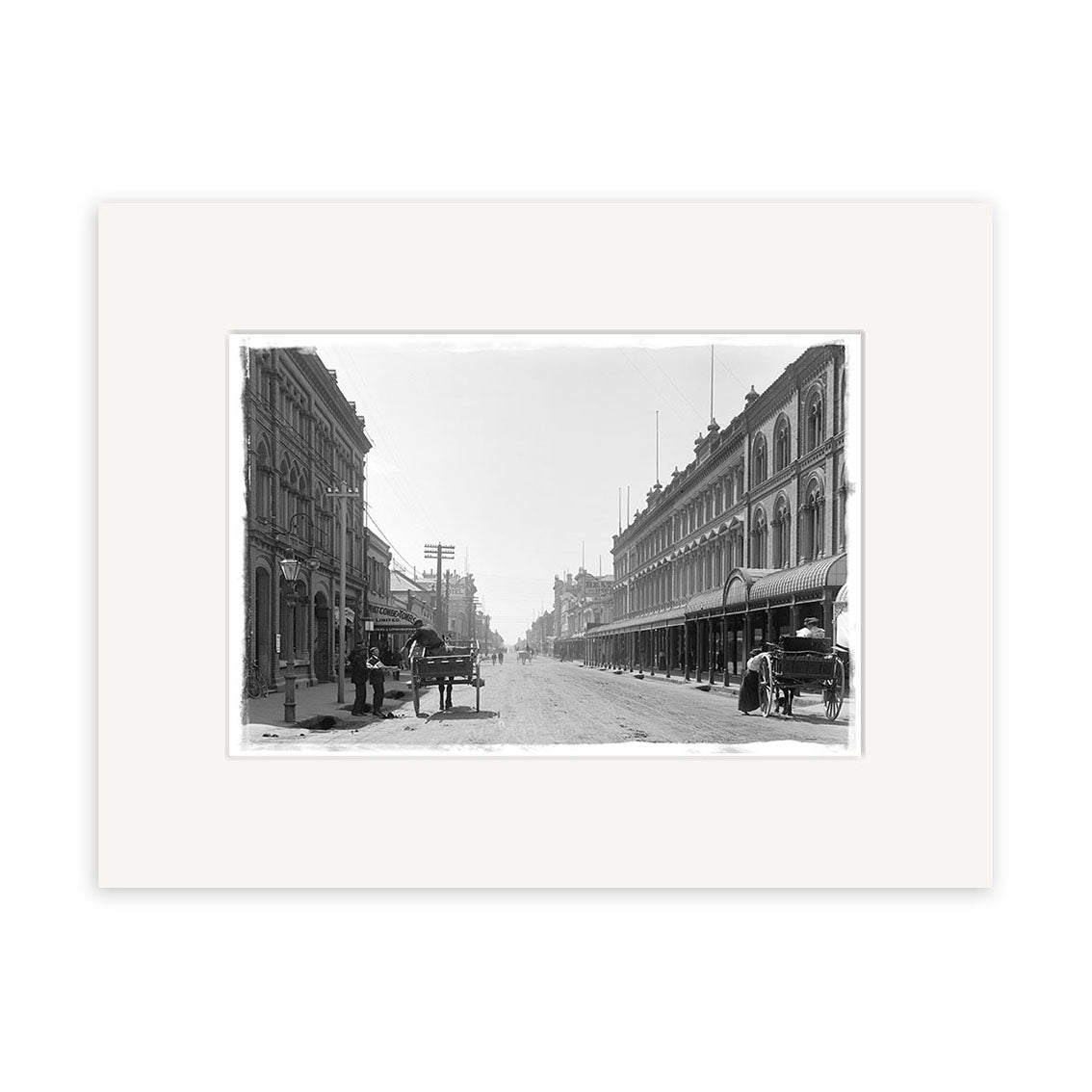 Archival print: Cashel St 1900 - HI025 shows a wide New Zealand street in the early 20th century, lined with multi-story buildings, horse-drawn carriages parked at the sides, and a few pedestrians.