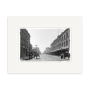 Archival print: Cashel St 1900 - HI025 shows a wide New Zealand street in the early 20th century, lined with multi-story buildings, horse-drawn carriages parked at the sides, and a few pedestrians.