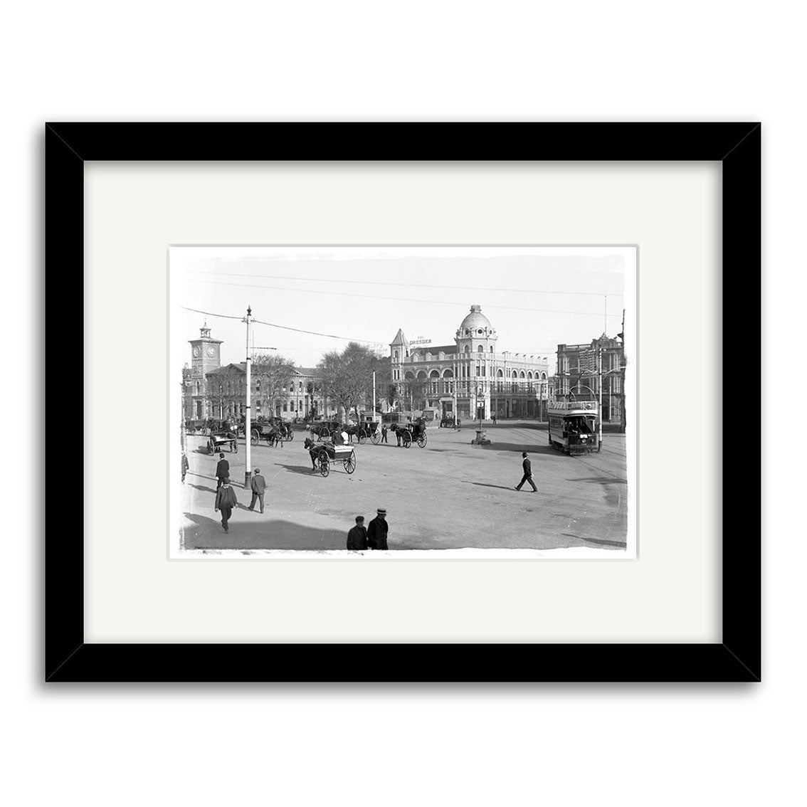 Cathedral Square 1910 - HI026: Black and white historic photographic print of a bustling New Zealand city street with people, horse-drawn carriages, early cars, and grand buildings, framed in a simple black border for archival quality.