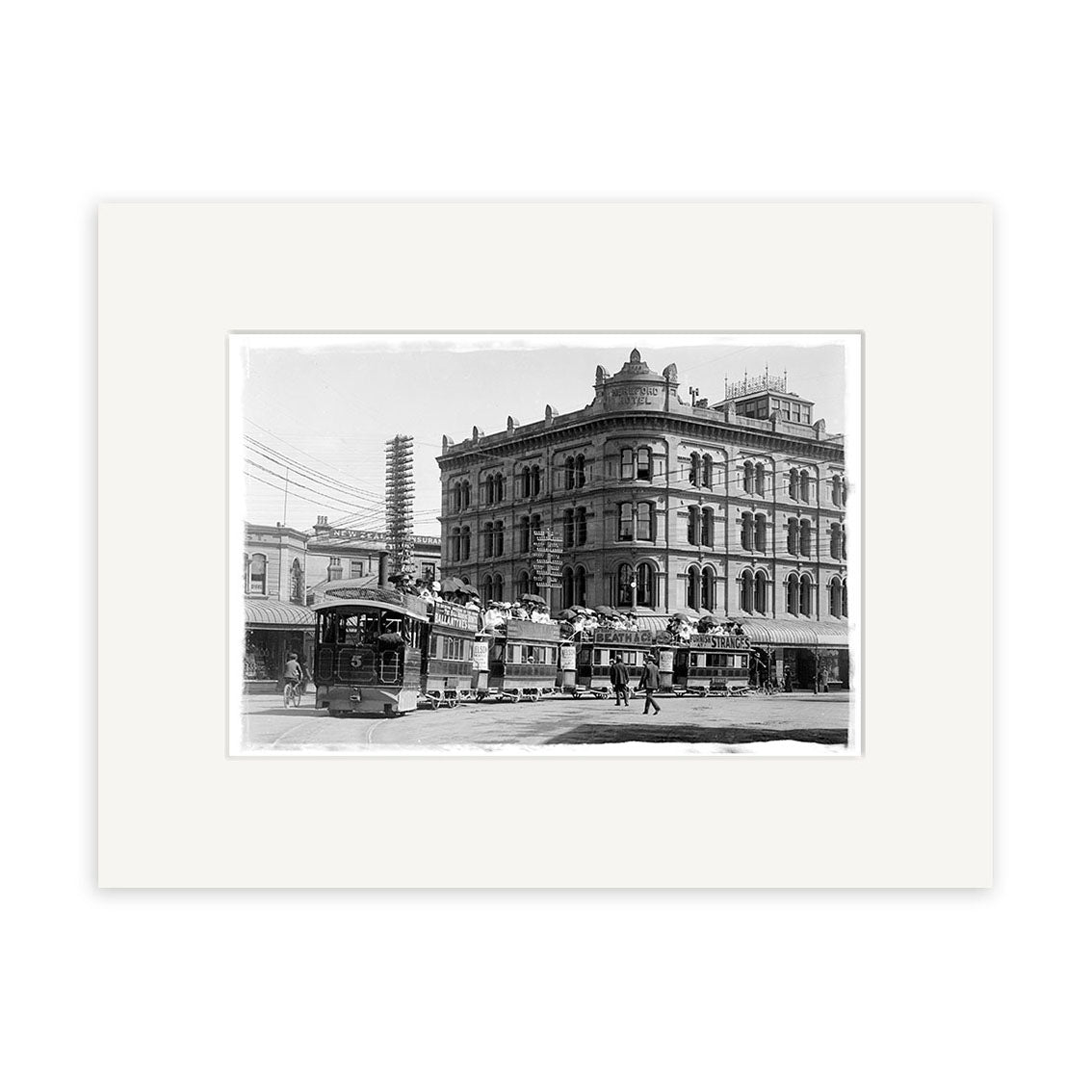 Black-and-white historical photographic print Puffing Billy 1902 - HI027 shows an old-fashioned streetcar and pedestrians by a grand building in New Zealand, with utility wires above. Print is on archival matte paper and framed with a white mat.