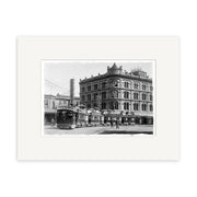 Black-and-white historical photographic print Puffing Billy 1902 - HI027 shows an old-fashioned streetcar and pedestrians by a grand building in New Zealand, with utility wires above. Print is on archival matte paper and framed with a white mat.