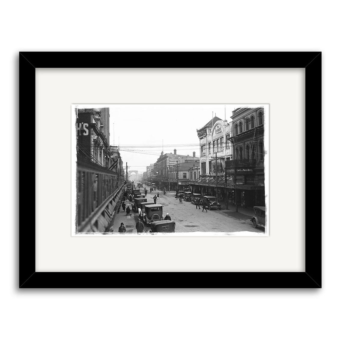 Cashel St 1930 - HI028: Black-and-white historical print of a New Zealand city street in the early 20th century featuring horse-drawn carriages, vintage cars, and pedestrians. Presented in a black frame with white mat.