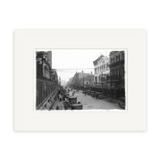 Archival-quality black-and-white photographic print: Cashel St 1930 - HI028 captures a bustling New Zealand city street with cars, horse-drawn wagons, and pedestrians among multi-story buildings from the early 20th century.