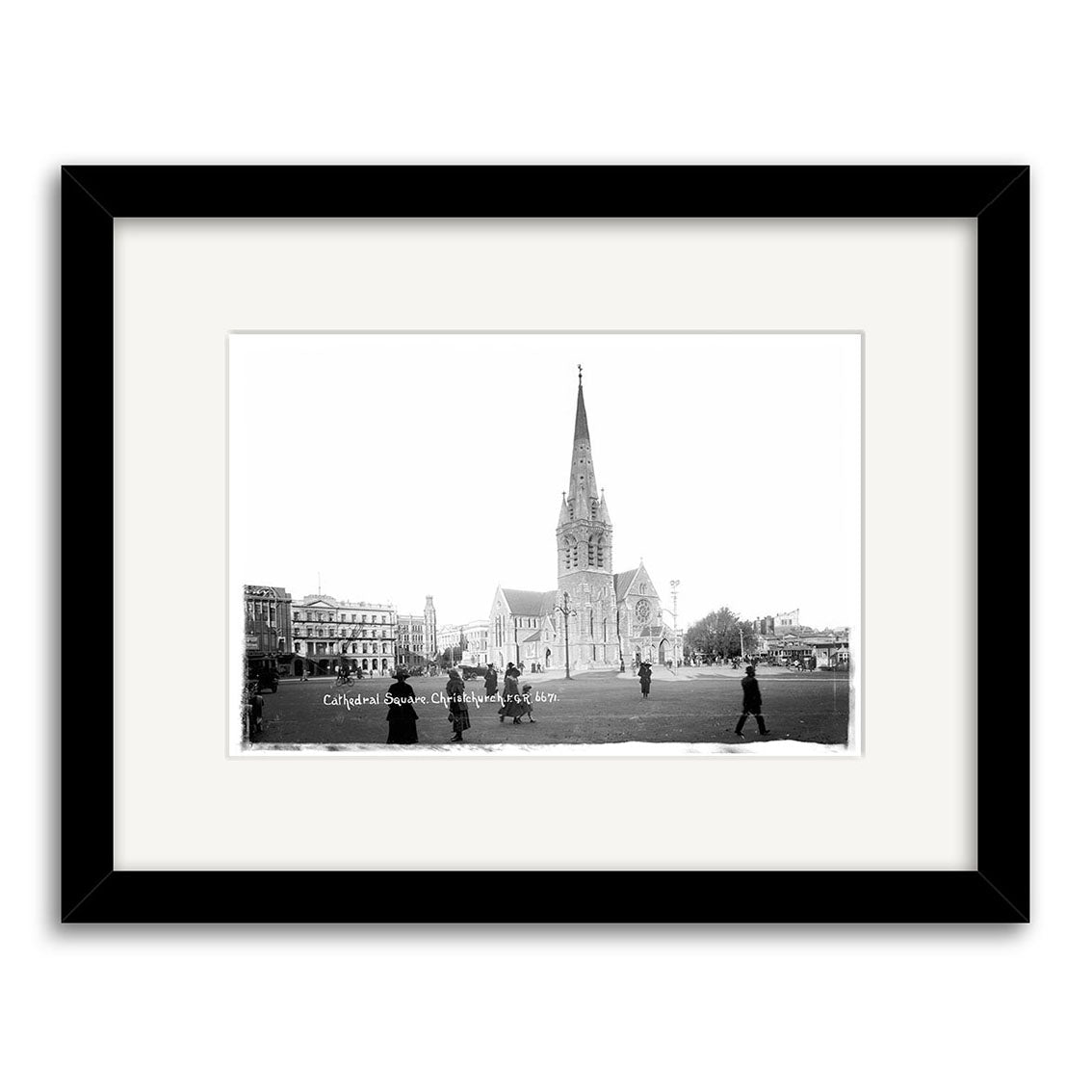 Black and white historical print of Christchurch Cathedral 1919 (HI029), featuring its tall central spire and nearby buildings. A few people walk in the open space. Framed with a white mat, black frame, and printed on archival quality matte paper.