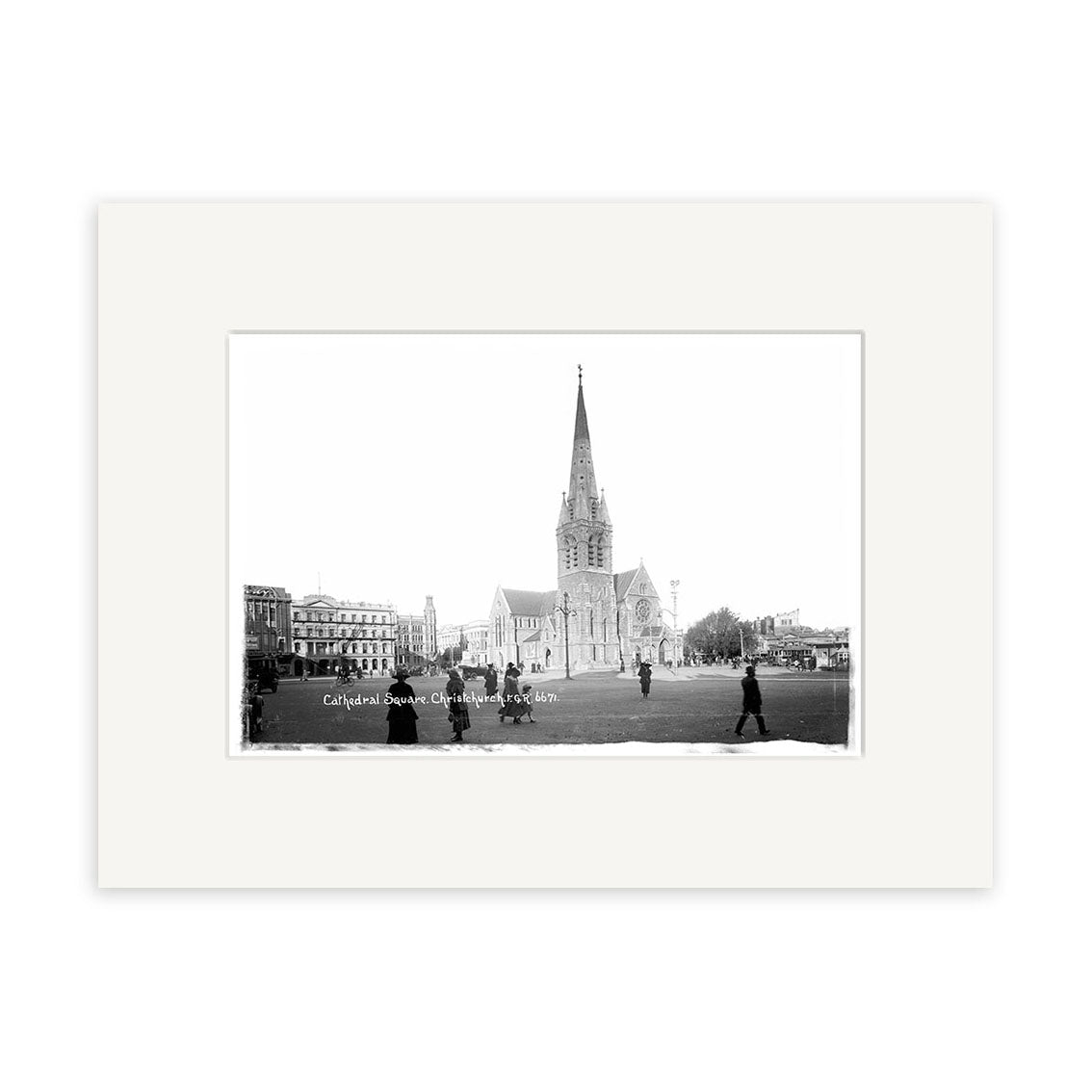 Black and white photo Christchurch Cathedral 1919 - HI029 features a tall-spired cathedral, surrounding grass, people walking in front, and nearby buildings, all presented in a white matte frame on archival quality matte paper.