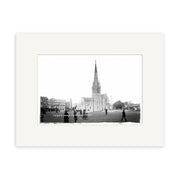 Black and white photo Christchurch Cathedral 1919 - HI029 features a tall-spired cathedral, surrounding grass, people walking in front, and nearby buildings, all presented in a white matte frame on archival quality matte paper.