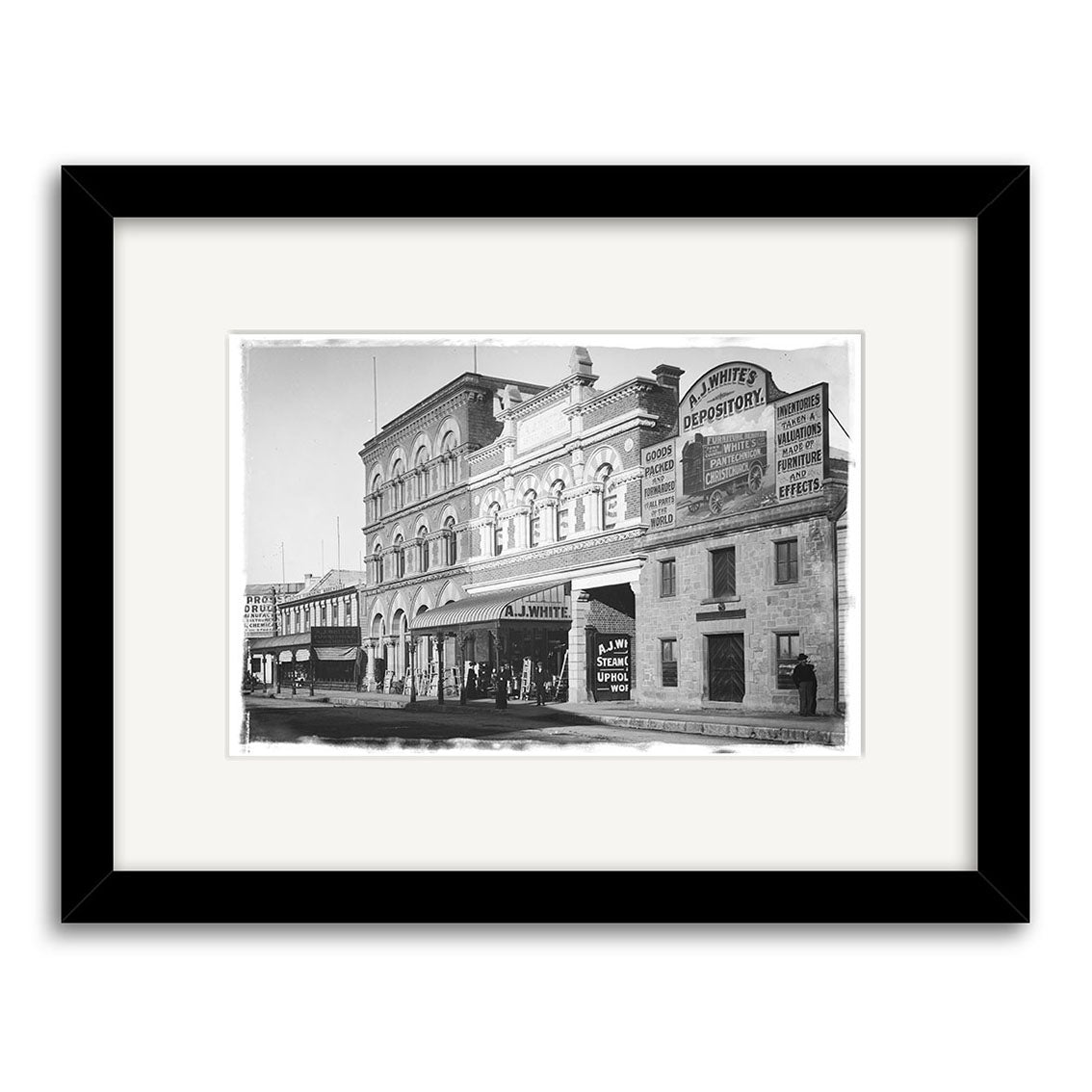 Black and white historical photographic print titled Tuam St 1900 - HI030 shows a New Zealand street with a large brick building, storefronts including White’s Depository sign, and people on the sidewalk. Framed in black for archival quality preservation.