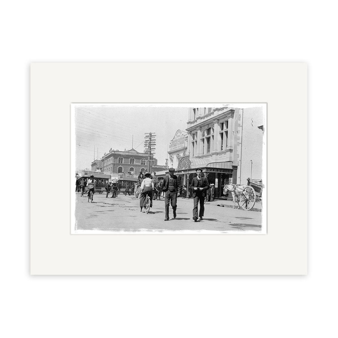 Black and white historical photographic print, Cnr High & Cashel St - HI032, shows a busy early 20th-century street scene with people, a horse-drawn carriage, and detailed buildings. Printed on archival quality matte paper for lasting clarity.