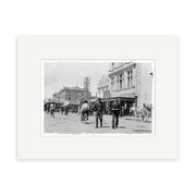 Black and white historical photographic print, Cnr High & Cashel St - HI032, shows a busy early 20th-century street scene with people, a horse-drawn carriage, and detailed buildings. Printed on archival quality matte paper for lasting clarity.