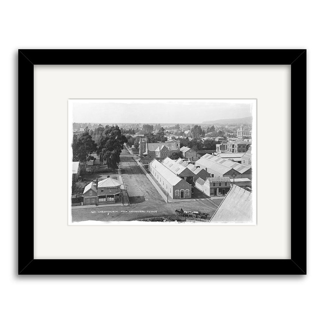 Black-and-white archival photographic print titled Worcester St 1882 - HI033, depicting a small New Zealand town with tree-lined streets, single-story buildings, and distant hills. Presented in a black frame with a white mat.