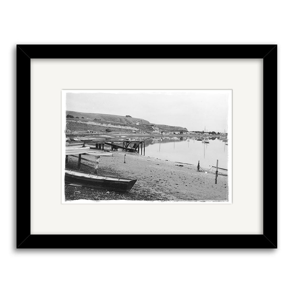 Moncks Bay 1914 - HI034: Black and white historical photographic print of a tranquil New Zealand waterfront with dock, small boat, calm water, and background buildings. Presented in an archival black frame with white mat.