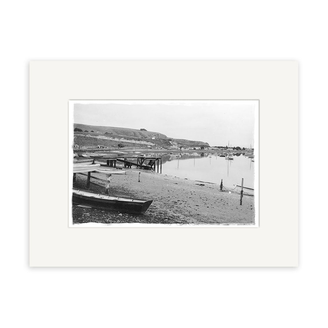 Moncks Bay 1914 - HI034: Black and white photo of a calm New Zealand waterfront with boats, docks, hills, buildings, and church spire. Historical print displayed with a white mat.