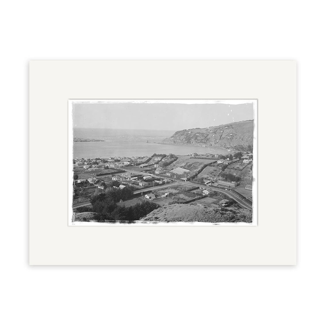 Redcliffs 1920 - HI035: A black and white photo of a New Zealand coastal town with houses, streets, and trees in front, water and hills behind. Printed on archival matte paper with a white border.