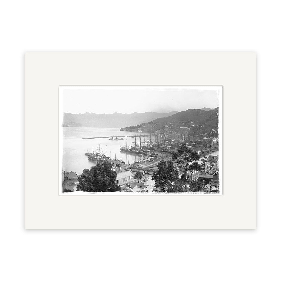 Lyttelton 1890 - HI036: Black and white photo of a New Zealand harbor with docked ships, buildings in the front, and hills beyond. Printed on archival quality matte paper and centered within a wide white mat.