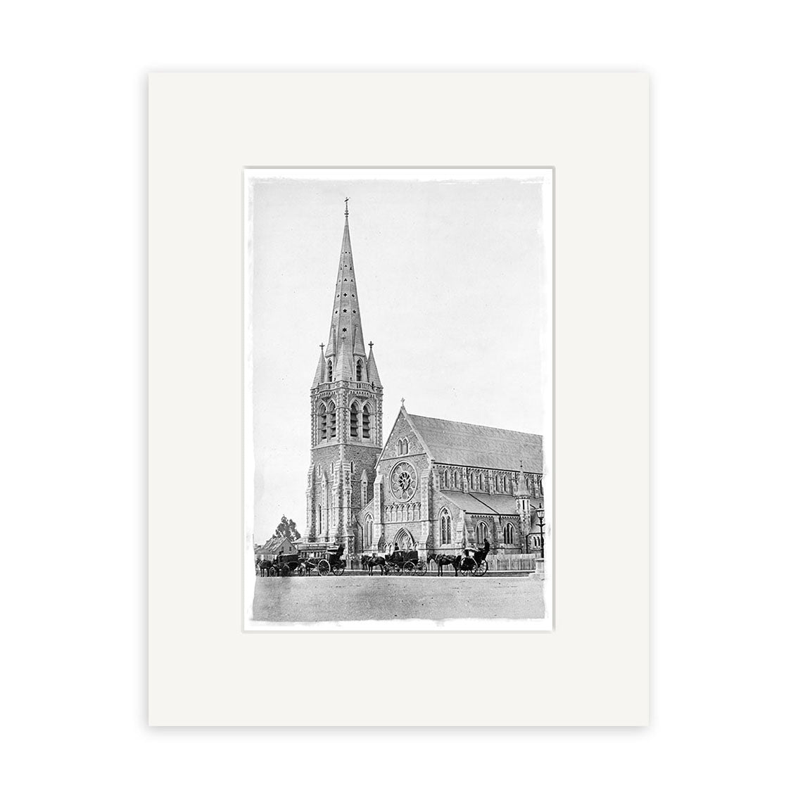 Black and white photo of Christchurch Cathedral 1880 (HI038), a large Gothic-style church with tall steeple and rose window. Horse-drawn carriages are parked in front. Historical print displayed with a wide white mat border.