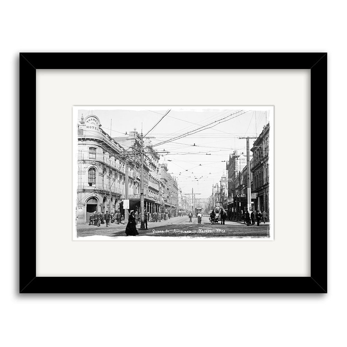 Archival quality black-and-white photographic print of Queen Street Auckland (HI042) features people, horse-drawn carriages, electric wires, multi-story buildings, and a hotel sign. Framed with a white mat.