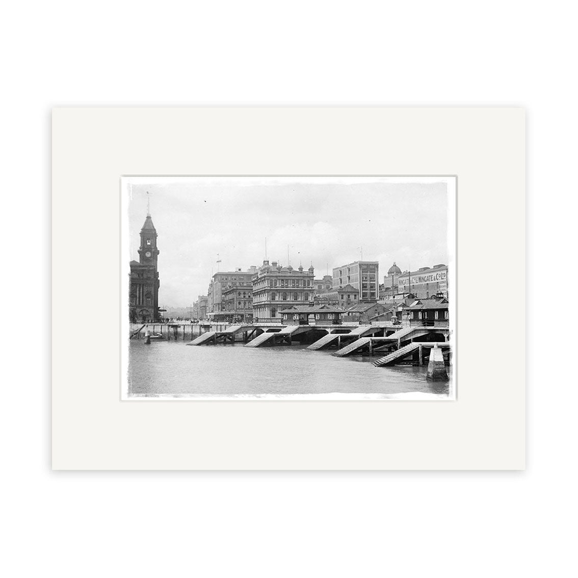 Archival-quality black and white photographic print, “Auckland Water Front 1916 - HI043,” shows a New Zealand cityscape with a river, bridge, large buildings, and a clock tower on the left, all bordered by a white matte.