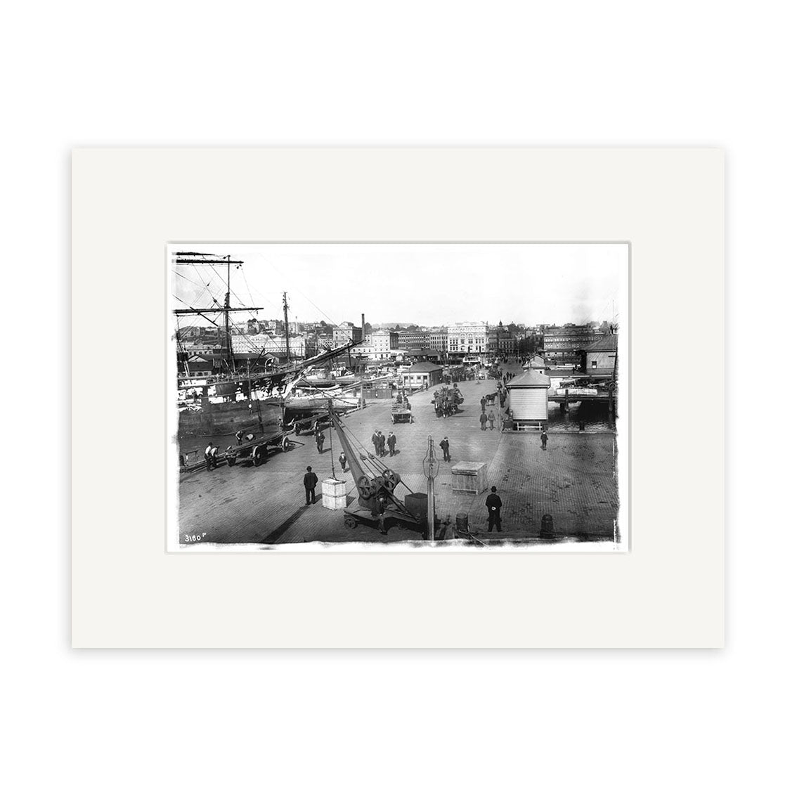 Queens Wharf 1905 - HI045: Black-and-white photograph showing a bustling New Zealand city street with pedestrians, horse-drawn carriages, early cars, buildings, and a crane in front. Presented in an archival quality white mat.