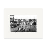 Queens Wharf 1905 - HI045: Black-and-white photograph showing a bustling New Zealand city street with pedestrians, horse-drawn carriages, early cars, buildings, and a crane in front. Presented in an archival quality white mat.