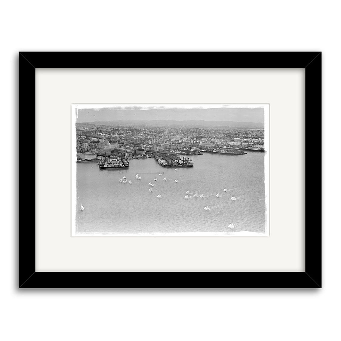 Anniversary Regatta 1950 - HI047: Black and white aerial photo of a New Zealand harbor with docks, large ships, and sailboats; this historical print is framed with a white mat and black frame.