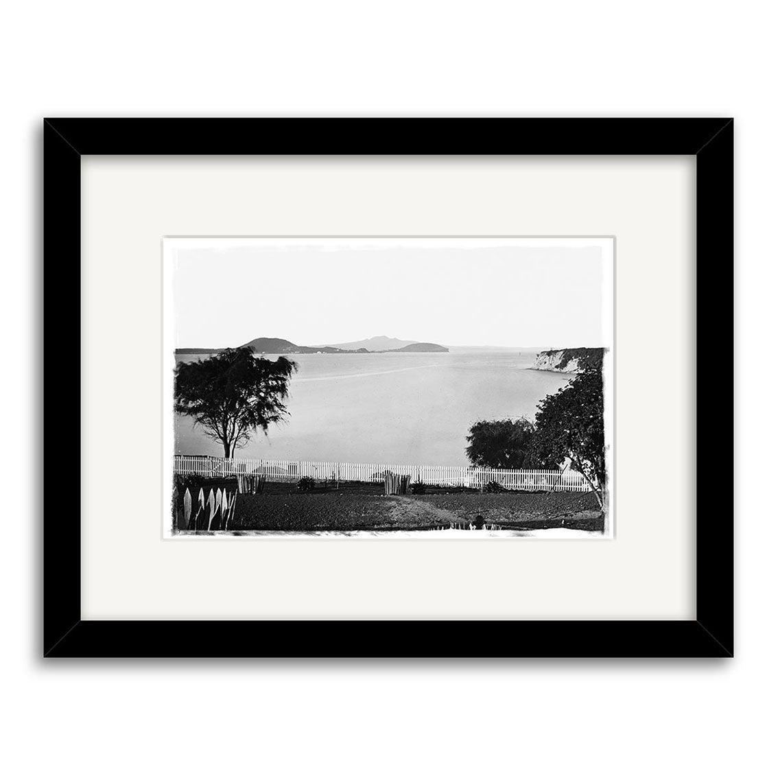 “St Georges Bay 1864 - HI049: Black and white photo of calm water with distant islands, trees, and fencing in the foreground. Printed on matte paper, framed in black with a white mat.”.